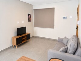 Uma sala de estar moderna com uma TV de tela plana em um suporte de madeira, um sofá cinza e uma grande janela com persiana bege.