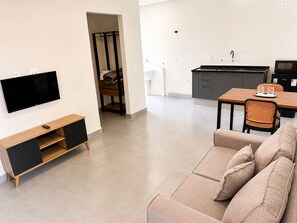 A modern living room with a flat-screen TV, a sofa, a dining table with chairs, and a kitchen area with a microwave and cabinets.