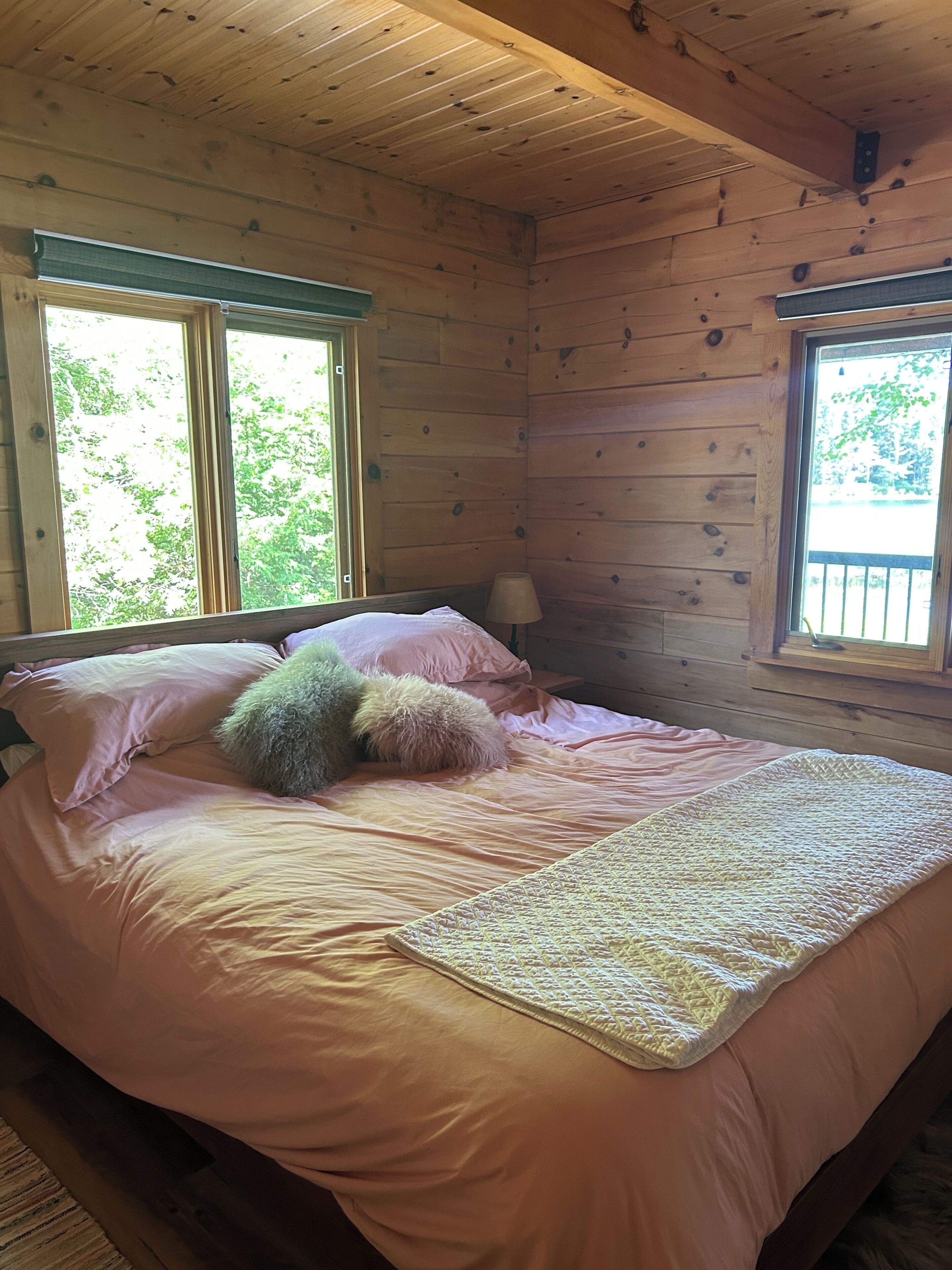 A wooden cabin bedroom with a bed, pillows, a lamp, and two windows.