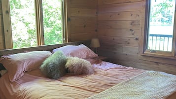 A wooden cabin bedroom with a bed, pillows, a lamp, and two windows.