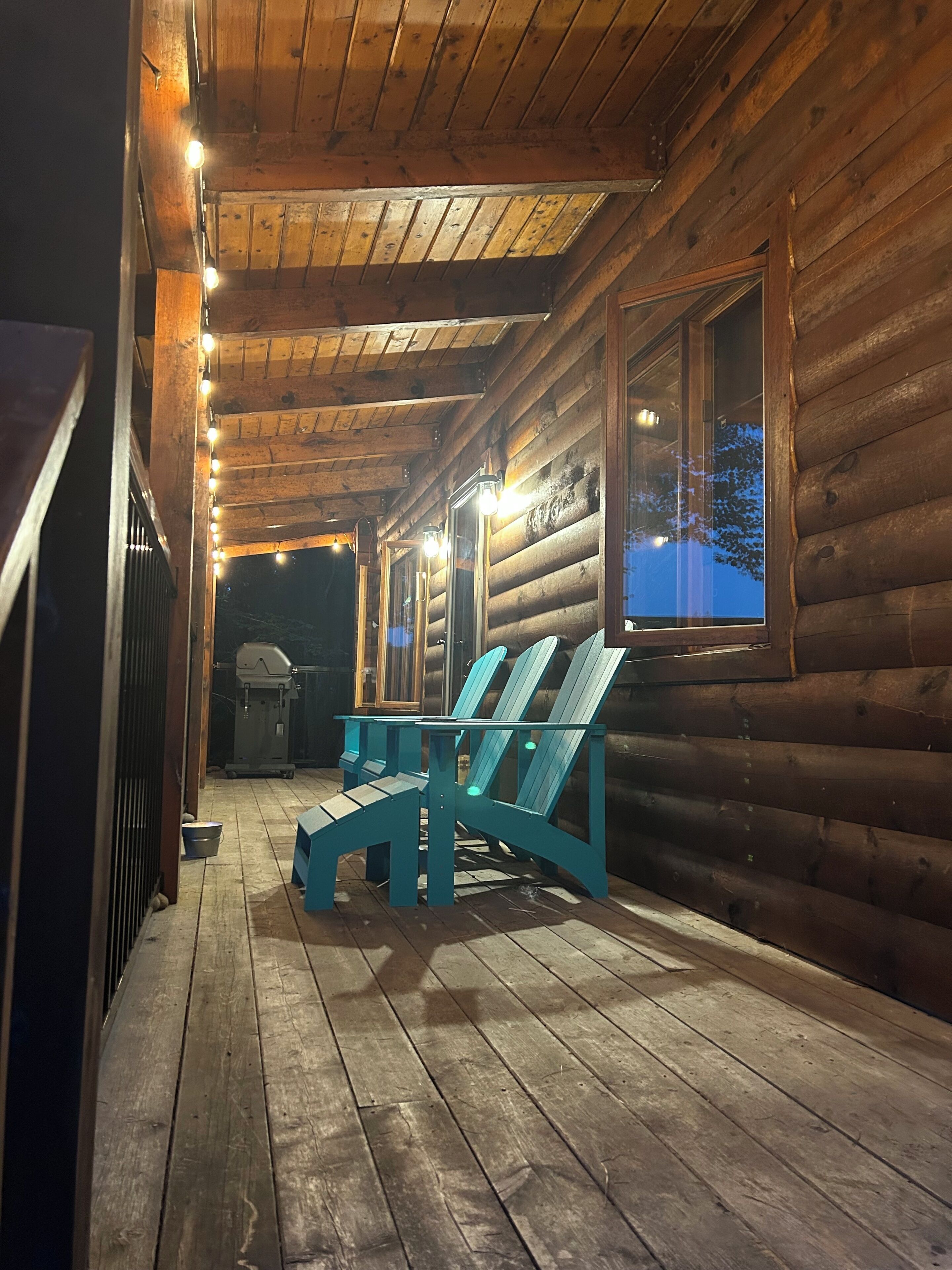A log cabin with a wooden porch, a grill, and two Adirondack chairs.