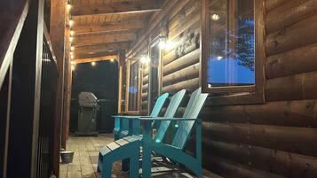 A log cabin with a wooden porch, a grill, and two Adirondack chairs.