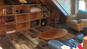 A cozy loft-style room with a flat-screen TV, bookshelves, a leather sofa, and a wooden floor.
