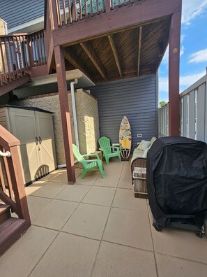 Ein überdachter Patio-Bereich mit Grill, zwei grünen Plastikstühlen, einem Surfbrett und einem abschließbaren Stauraum.