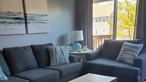 A modern living room with a grey sectional sofa, a wooden coffee table, and a view of a house through a large window. - Coastal Comfort - 2BR/2BA Condo in North OCMD - Walk to Beach, Dining & Fun (Ocean City)
