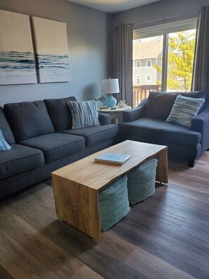 Ett modernt vardagsrum med en grå hörnsoffa, ett soffbord i trä och utsikt över ett hus genom ett stort fönster.