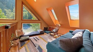 A cozy attic bedroom with a sloped ceiling, large windows, a blue sofa, and a rocking chair. - Riverfront A-Frame with Hot Tub, Game Room and Fire Pit - Sleeps 10, Dogs OK (Granite Falls)