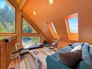 A cozy attic bedroom with a sloped ceiling, large windows, a blue sofa, and a rocking chair. - Riverfront A-Frame with Hot Tub, Game Room and Fire Pit - Sleeps 10, Dogs OK (Granite Falls)