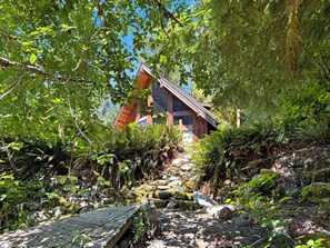 A wooden cabin with a sloped roof, surrounded by dense greenery and a stone pathway leading to it. - Riverfront A-Frame with Hot Tub, Game Room and Fire Pit - Sleeps 10, Dogs OK (Granite Falls)