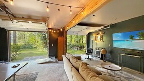 A modern living room with a large sofa, a television, and a view of a garden. - Riverfront A-Frame with Hot Tub, Game Room and Fire Pit - Sleeps 10, Dogs OK (Granite Falls)
