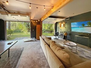 A modern living room with a large sofa, a television, and a view of a garden. - Riverfront A-Frame with Hot Tub, Game Room and Fire Pit - Sleeps 10, Dogs OK (Granite Falls)