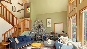 A cozy living room with a stone fireplace, a blue sofa, a wooden staircase, and a large window. - Riverfront A-Frame with Hot Tub, Game Room and Fire Pit - Sleeps 10, Dogs OK (Granite Falls)