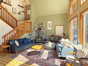 A cozy living room with a stone fireplace, a blue sofa, a wooden staircase, and a large window. - Riverfront A-Frame with Hot Tub, Game Room and Fire Pit - Sleeps 10, Dogs OK (Granite Falls)