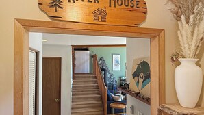 A wooden sign with 'River House' and a house icon, a staircase leading to a living area with a couch and a painting, and a white vase with pampas grass. - Riverfront A-Frame with Hot Tub, Game Room and Fire Pit - Sleeps 10, Dogs OK (Granite Falls)