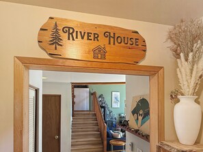 A wooden sign with 'River House' and a house icon, a staircase leading to a living area with a couch and a painting, and a white vase with pampas grass. - Riverfront A-Frame with Hot Tub, Game Room and Fire Pit - Sleeps 10, Dogs OK (Granite Falls)