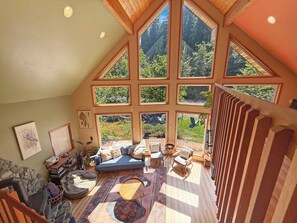 A spacious loft with a high ceiling, large windows offering a view of a forested area, a cozy seating area with a sofa and chairs, and a wooden staircase. - Riverfront A-Frame with Hot Tub, Game Room and Fire Pit - Sleeps 10, Dogs OK (Granite Falls)