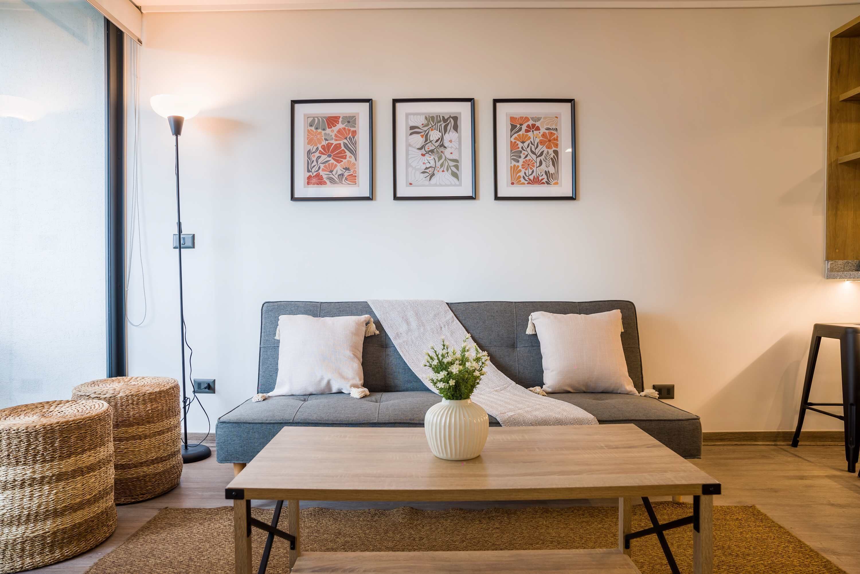 A modern living room with a grey sofa, two white pillows, a wooden coffee table with a white vase, and three framed floral artworks on the wall.