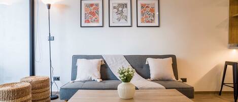 A modern living room with a grey sofa, two white pillows, a wooden coffee table with a white vase, and three framed floral artworks on the wall.