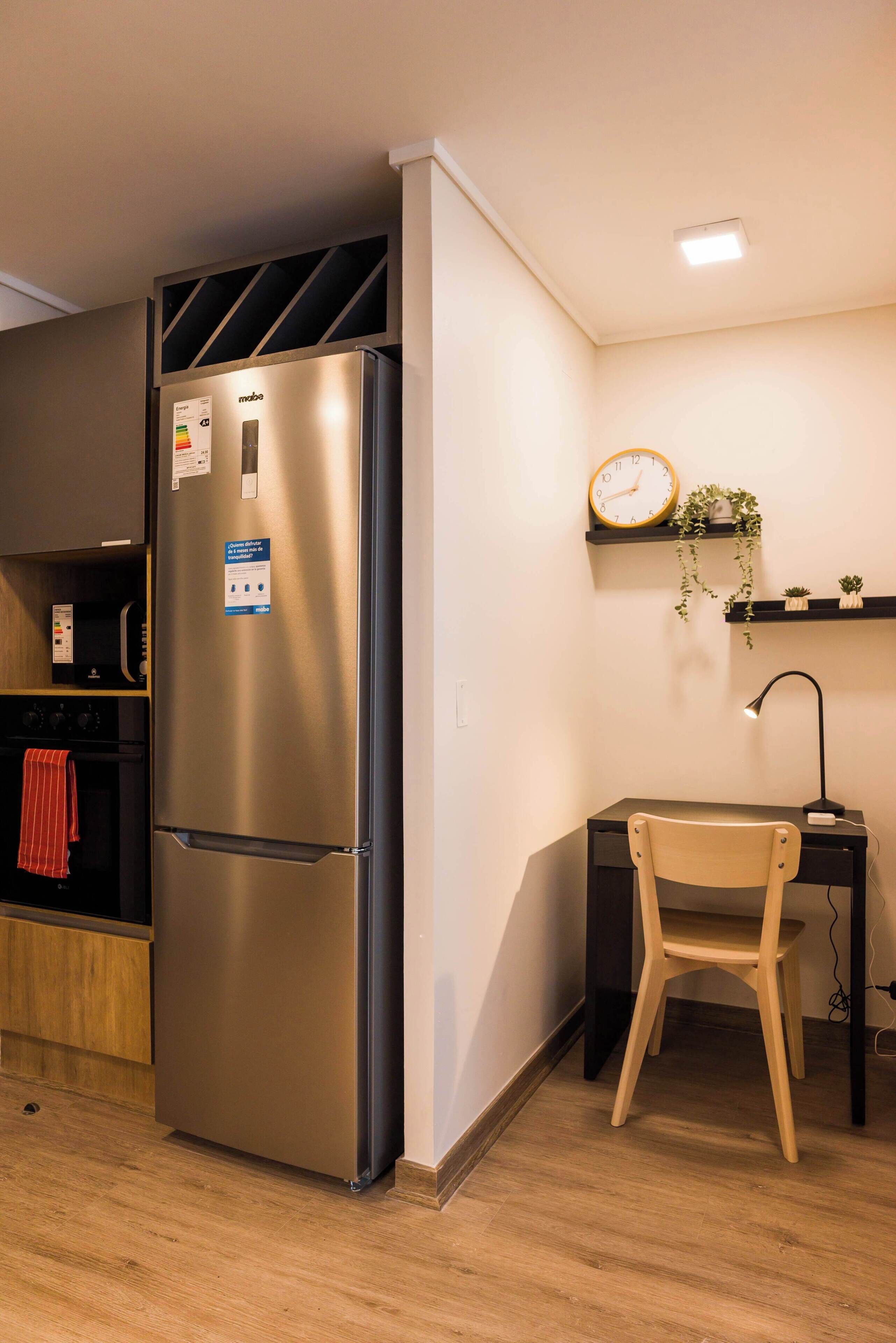 A compact kitchen with a stainless steel refrigerator, a wooden chair, a small desk with a lamp, and a wall-mounted clock.