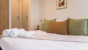 En pænt redt seng med sammenrullede hvide håndklæder, et natbord med en lampe og et indrammet billede på væggen.