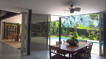 A dining area with a table and chairs, a ceiling fan, and a view of a swimming pool and garden through large glass doors.