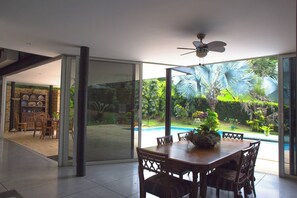 Uma área de refeições com mesa e cadeiras, ventilador de teto e vista para uma piscina e jardim através de grandes portas de vidro.
