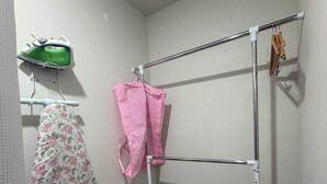Una esquina de una habitación con un tendedero de metal, un cesto blanco de ropa y una prenda con estampado floral colgada.