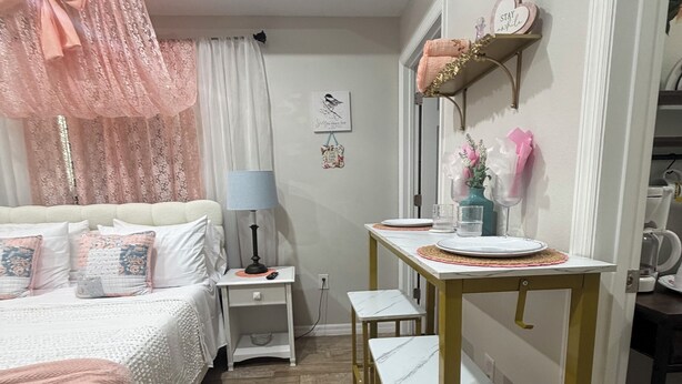 Un dormitorio con cama, mesitas de noche, un escritorio con sillas, una lámpara y una estantería con objetos decorativos.