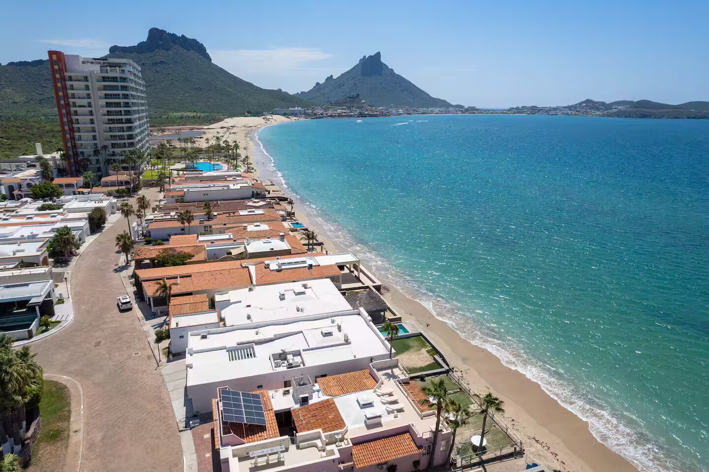 A coastal hotel with a beach view, multiple buildings, and a prominent mountain in the background.