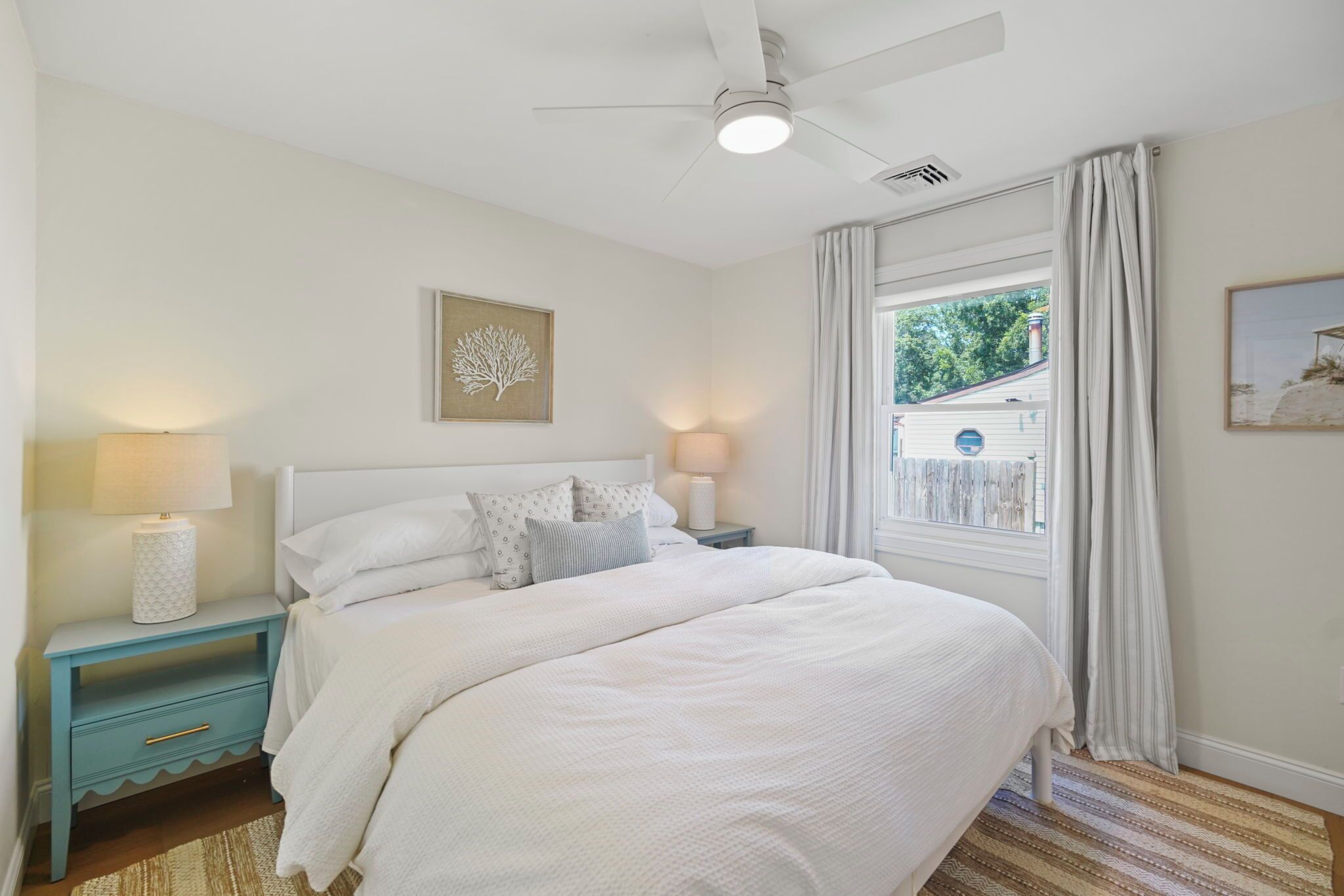A bedroom with a bed, nightstands, a ceiling fan, a window with curtains, and a wall-mounted picture.