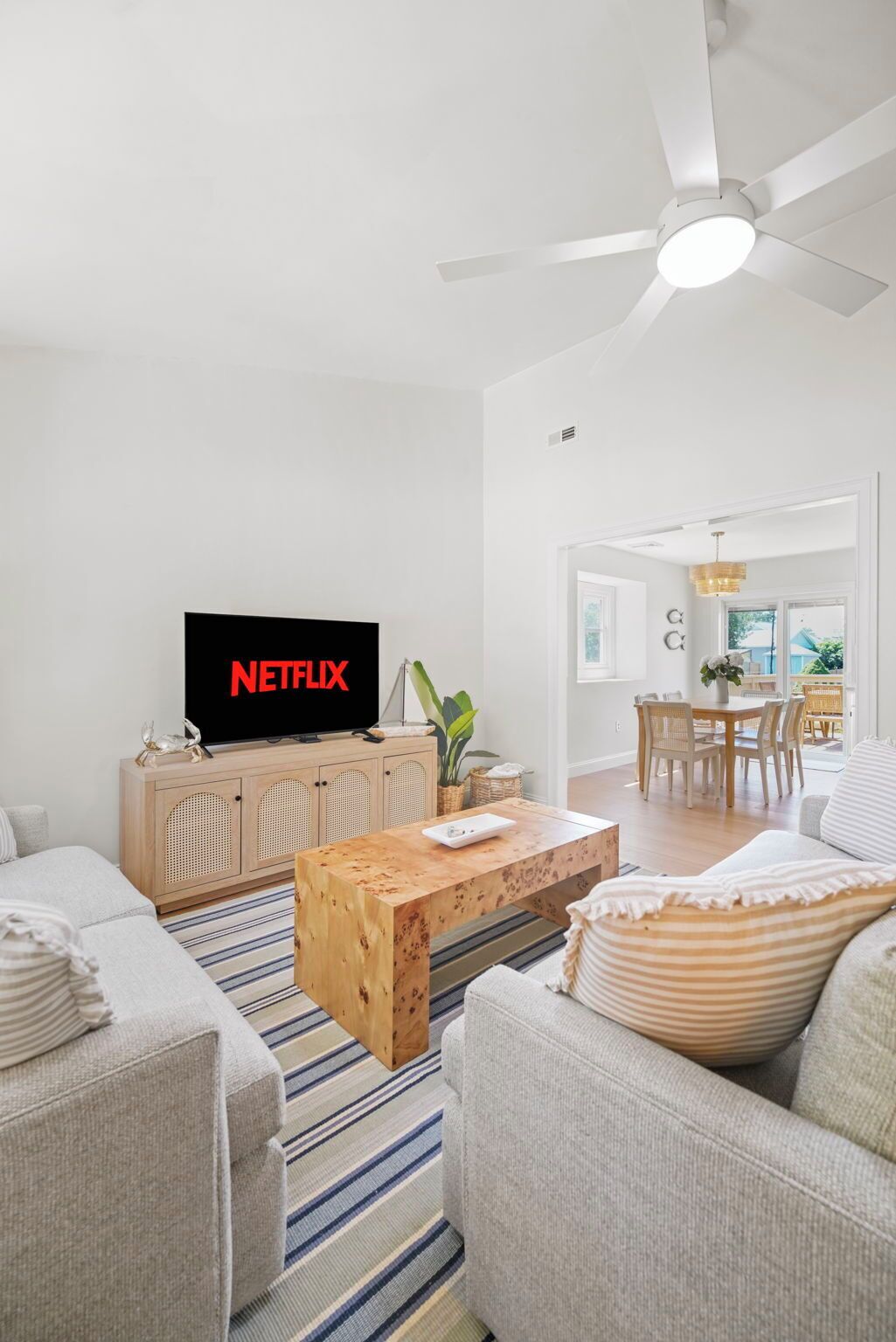 Ein modernes Wohnzimmer mit einem Flachbildfernseher, der 'NETFLIX' anzeigt, einem Holztisch, einem Streifenmuster-Teppich und einem Deckenventilator.