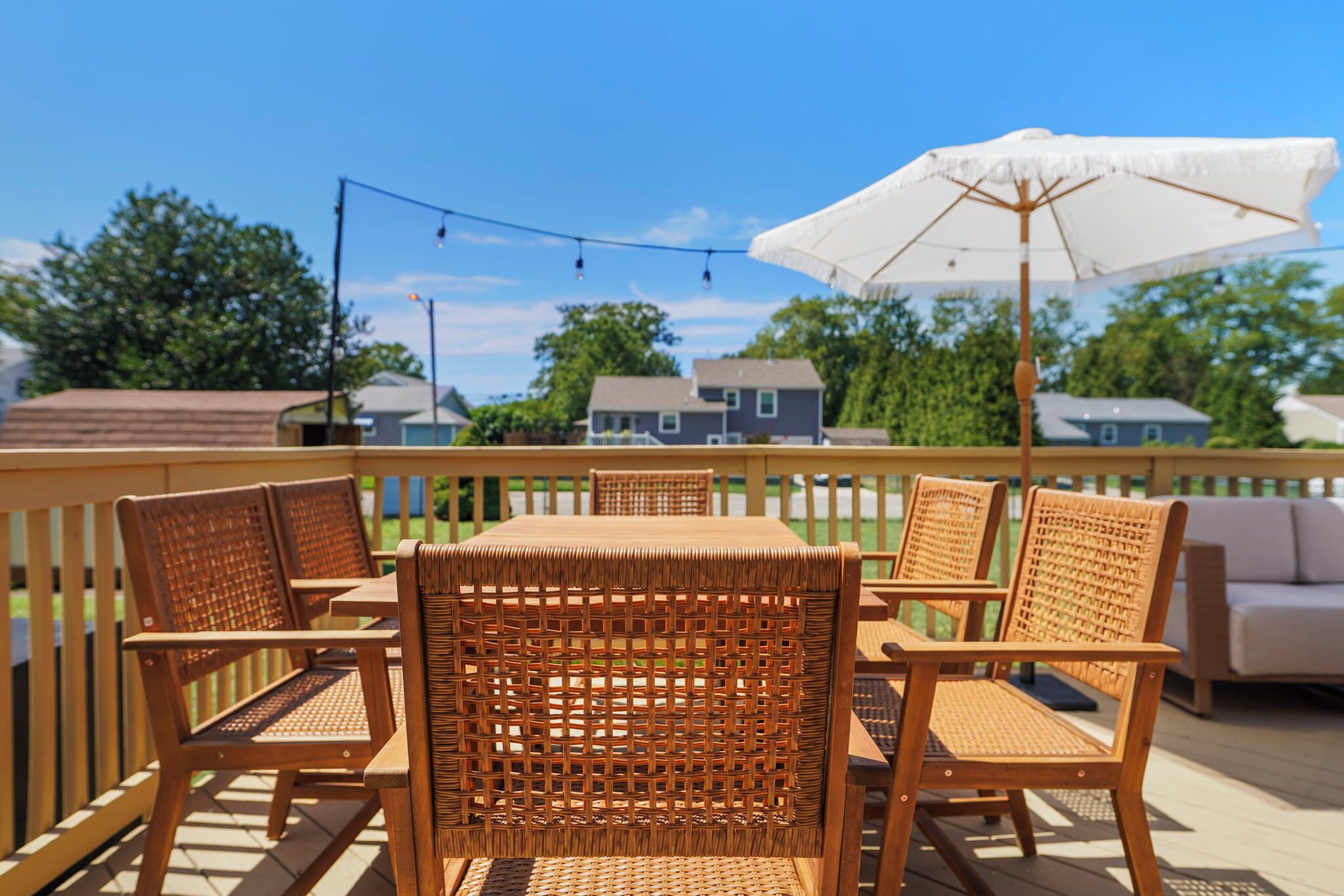 A deck with a wooden table and wicker chairs, an umbrella, and a view of houses in the background.