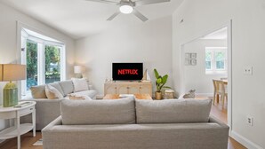 En stue med en grå sofa, et fjernsyn der viser 'NETFLIX', en loftventilator og udsigt til det fri gennem store vinduer.
