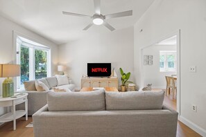 Una sala de estar con un sofá gris, un televisor mostrando 'NETFLIX', un ventilador de techo y vistas al exterior a través de ventanas grandes.