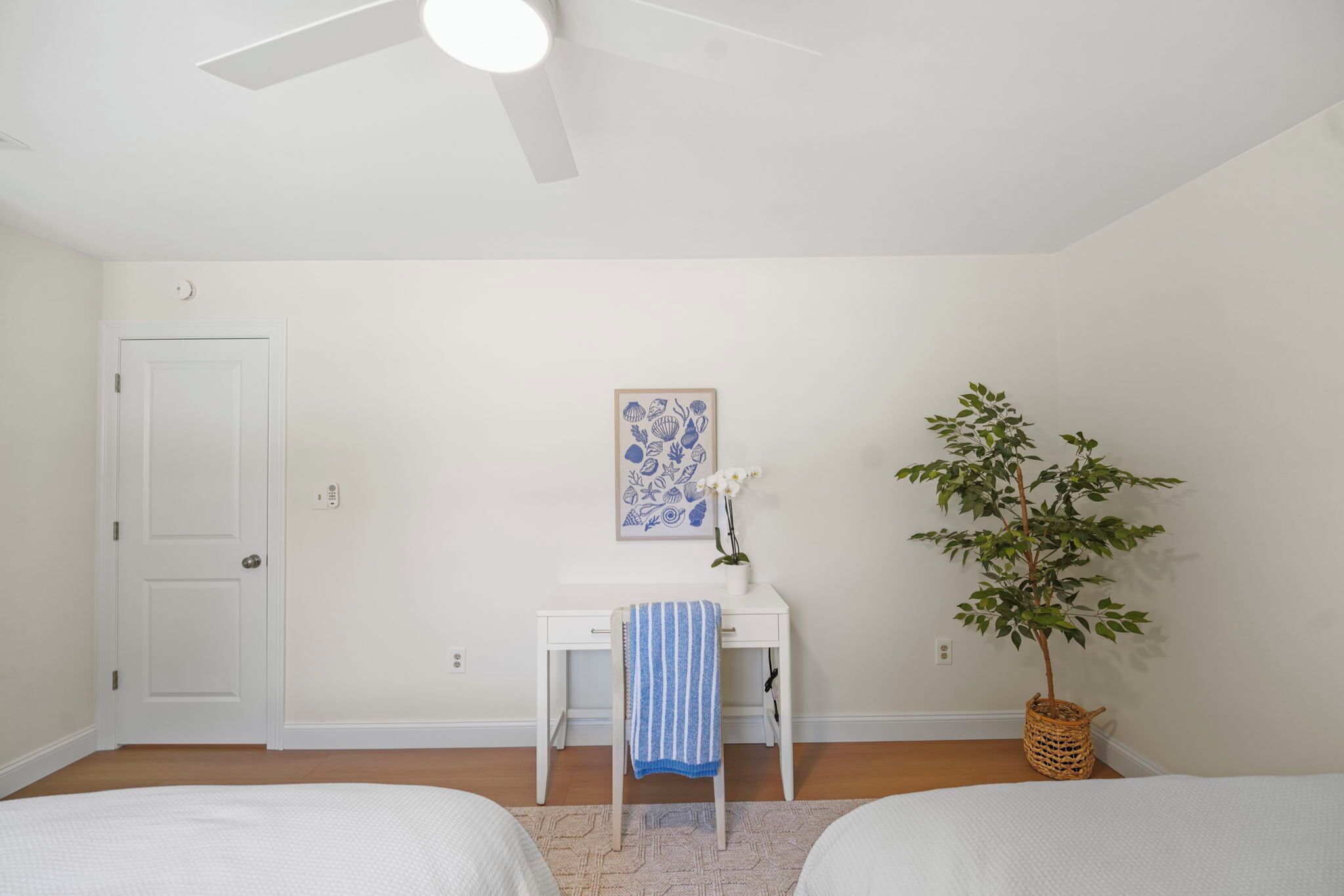 Une chambre avec deux lits, un bureau avec une chaise, un ventilateur de plafond, un tableau encadré au mur et une plante en pot.