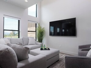 A modern living room with a sectional sofa, a flat-screen TV mounted on the wall, and a large window providing a view of the outdoors. - Stone Peak Mountain Retreat - New w/ ac (Driggs)