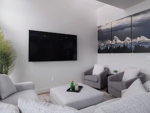 A modern living room with a flat-screen TV, grey sofas, a coffee table with a tray, and a large mountain landscape painting. - Stone Peak Mountain Retreat - New w/ ac (Driggs)