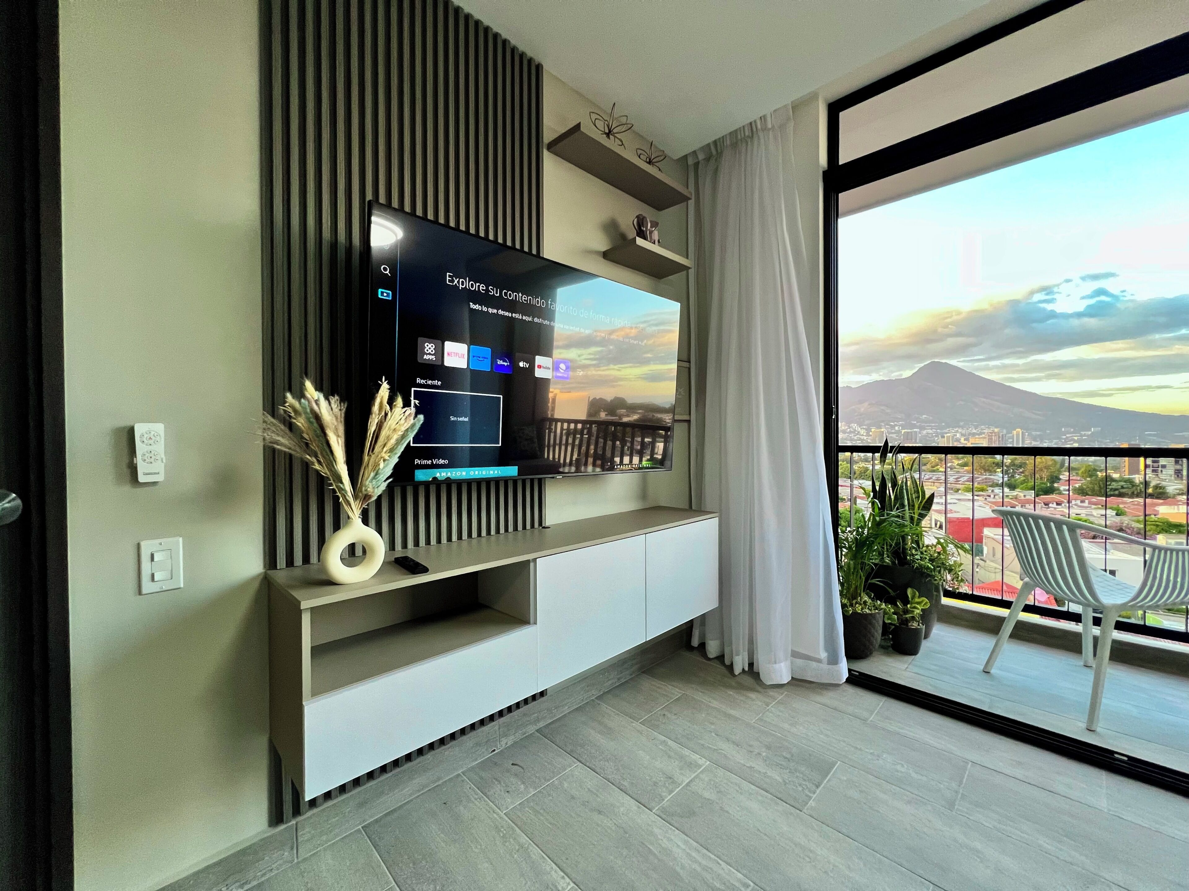 A modern living room with a flat-screen TV mounted on a wall, a balcony with a view of a cityscape and a mountain, and a minimalist design with a vase of dried plants on the console table.