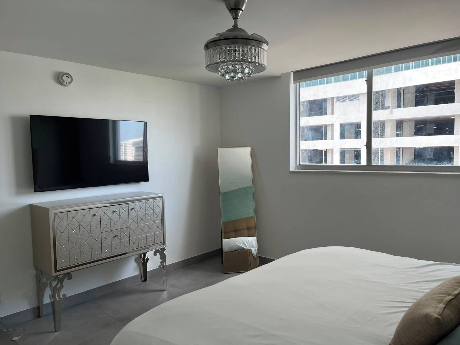 A bedroom with a bed, a TV, a dresser, a mirror, and a window with a view of buildings.