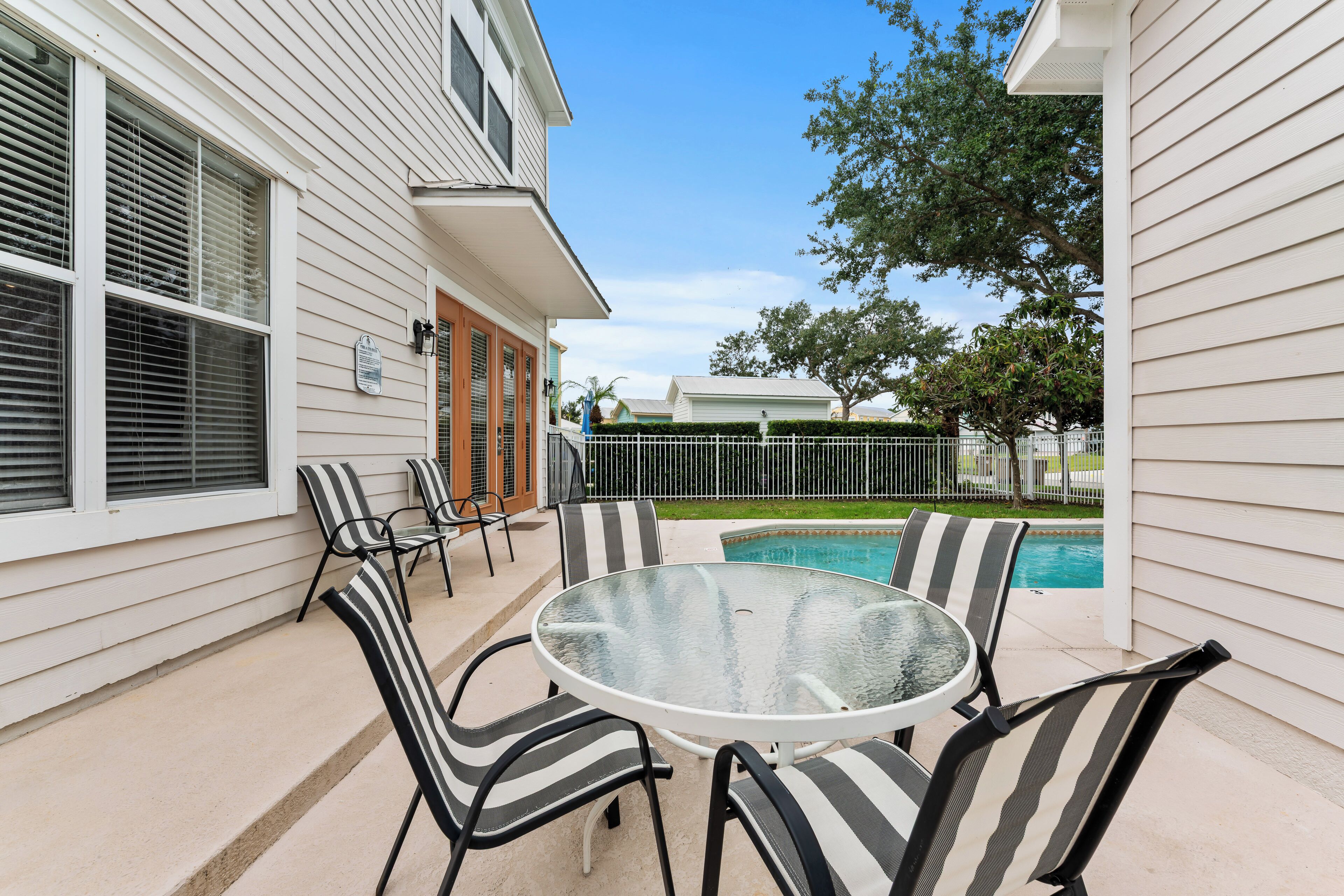 A patio with a glass-top table and striped chairs, a swimming pool, and a white house with shutters.