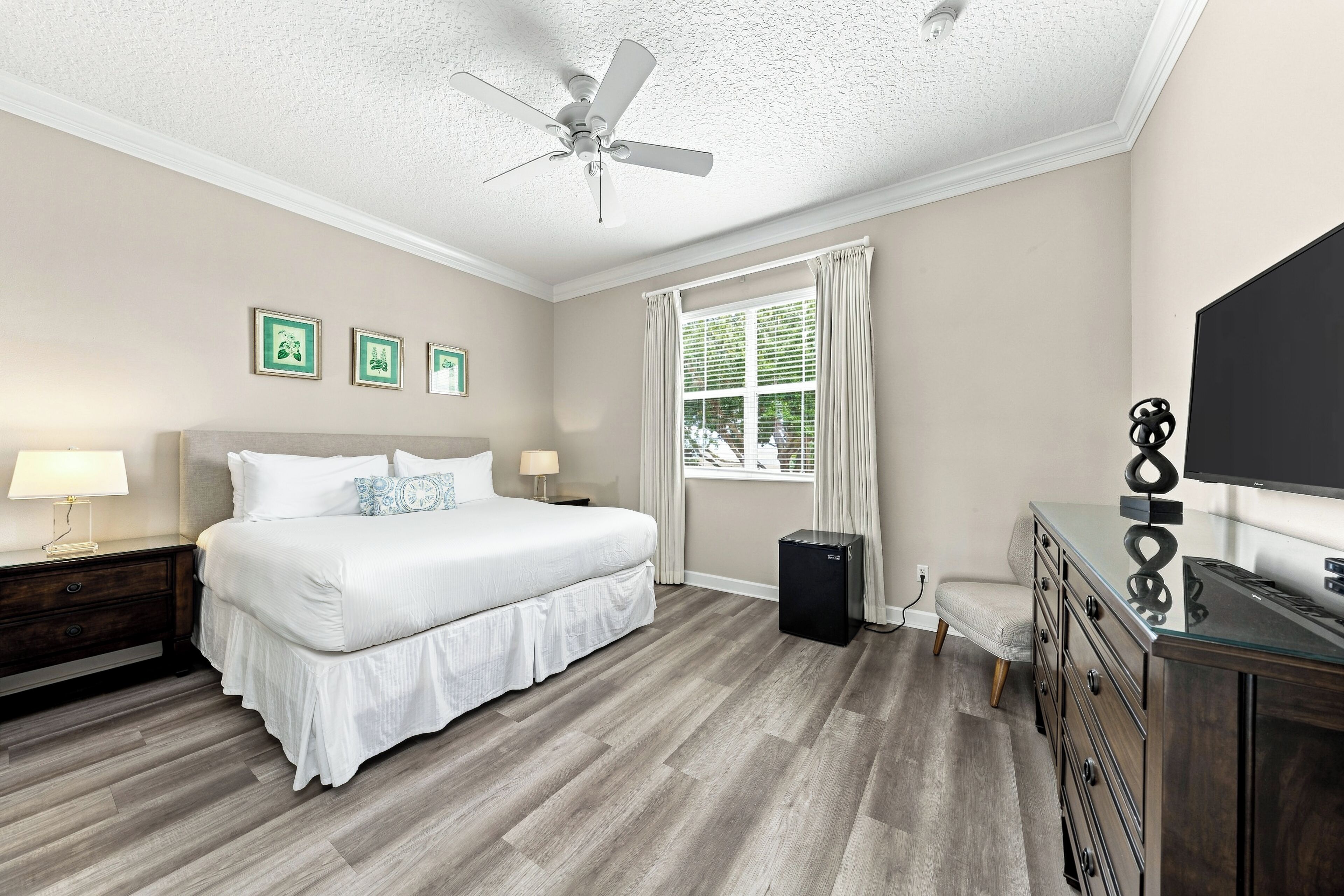 A bedroom with a bed, bedside tables, a ceiling fan, a window with curtains, and a television on a dresser.