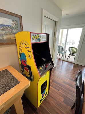 Una máquina arcade de Pac-Man en una habitación con suelo de madera, una puerta que da a un balcón y un cuadro colgado en la pared.