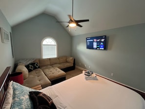 Un dormitorio con cama, sofá seccional, ventilador de techo, televisor montado en la pared y ventana con persianas.