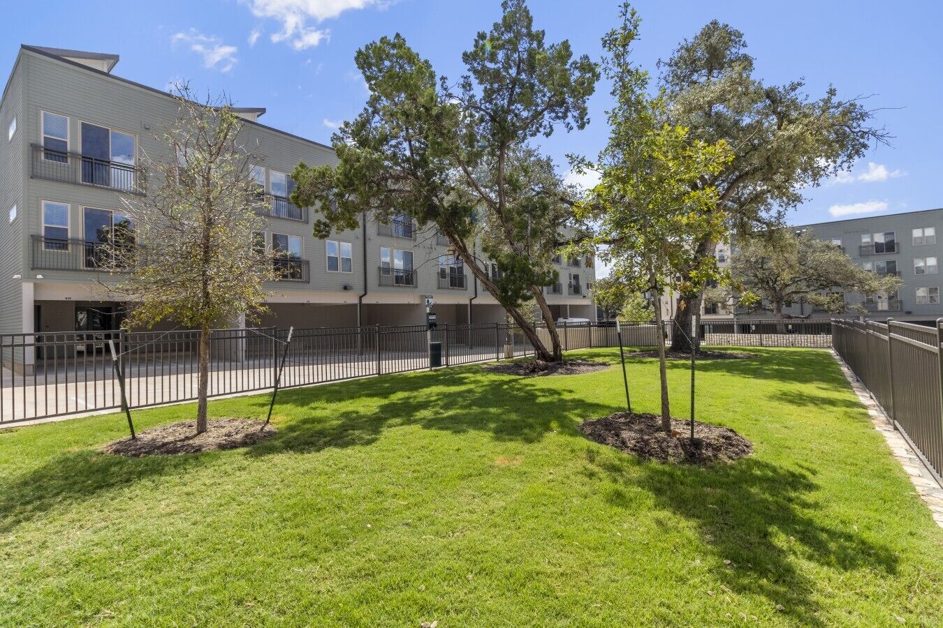 A residential building with multiple balconies, a fenced yard with young trees, and a clear sky.