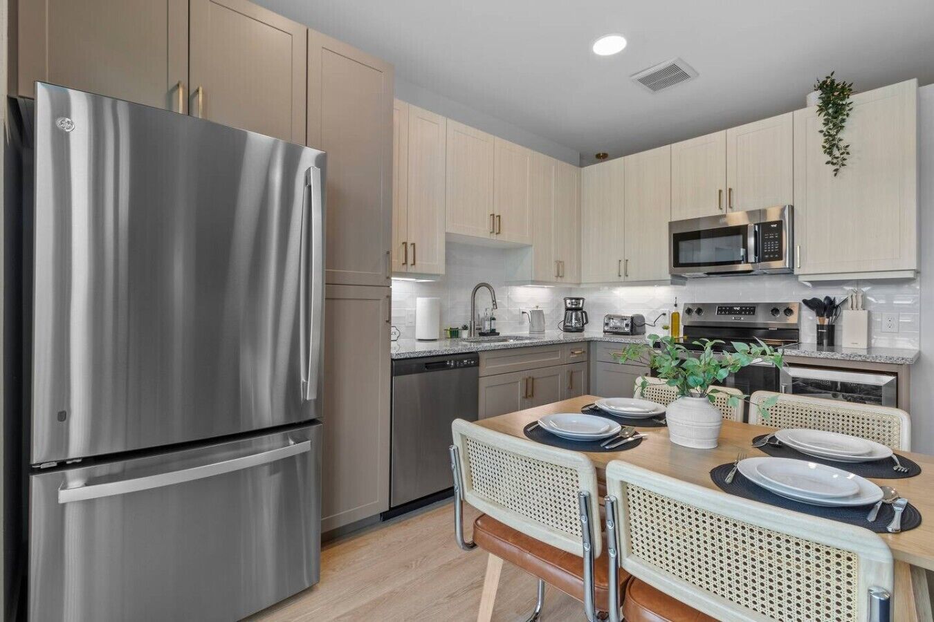 A modern kitchen with stainless steel appliances, light wood cabinetry, and a dining area with rattan chairs.