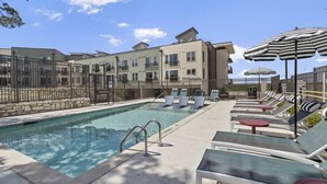 Une piscine entourée d’une clôture et de bâtiments, avec des chaises longues et des parasols à proximité.