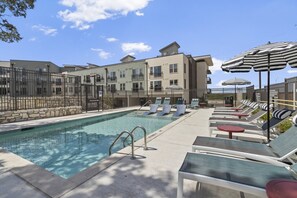 A swimming pool with lounge chairs and umbrellas, surrounded by a gated fence and buildings. - Stylish 1 King BR suite-Domain Gym, Pool & Parking (Austin)