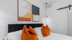 Une chambre d’hôtel avec un lit, du linge de lit blanc et des oreillers orange. Les murs sont ornés de tableaux abstraits et le plafond est éclairé par un luminaire.