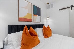 A hotel room with a bed, white bedding, and orange pillows. There are abstract paintings on the wall and a ceiling light fixture. - Stylish 1 King BR suite-Domain Gym, Pool & Parking (Austin)