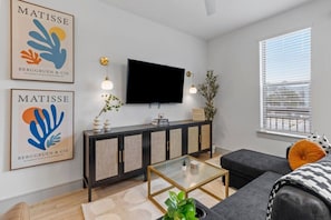 A modern living room with a flat-screen TV, a dark-colored sofa, a glass coffee table, and two wall-mounted artworks. - Stylish 1 King BR suite-Domain Gym, Pool & Parking (Austin)